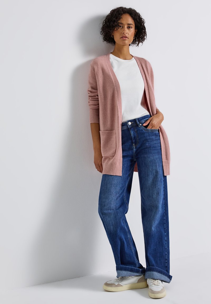 Cardigan rose clair avec deux poches avant, associé à un t-shirt blanc et un jean bleu foncé à jambes larges, porté avec des baskets crème.