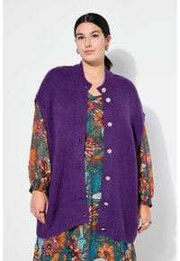 Lila gestrickte Cardigan mit Knopfleiste, im lockeren Schnitt und geripptem Saum, über einem bunten, floral bedruckten Kleid getragen.