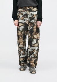 Άτομο με φαρδιά παντελόνια με αφηρημένο μοτίβο σε καφέ, μαύρο και γκρι χρώμα, μαύρο μπλουζάκι με μακριά μανίκια και γκρι αθλητικά παπούτσια, που στέκεται ακίνητο.