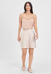 Camisola rosa clara com borda de renda, combinada com calções plissados bege. O modelo usa sapatilhas com perfurações, posando contra um fundo branco.