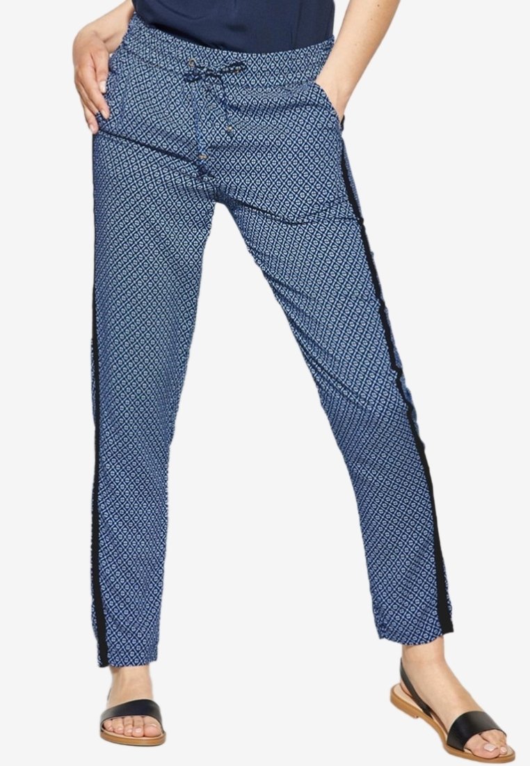 Femme portant un pantalon bleu à motifs avec une bande latérale noire, un haut noir et des sandales plates noires à bout ouvert, debout les mains dans les poches.