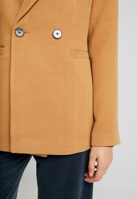 Kamelfärgad kavaj med en texturerad yta, dubbelknäppt design, två svarta och silverfärgade knappar samt sidofickor.