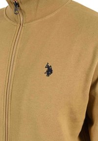 Light brown zip-up sweater with a ribbed collar and a small dark embroidered polo player logo on the left chest.