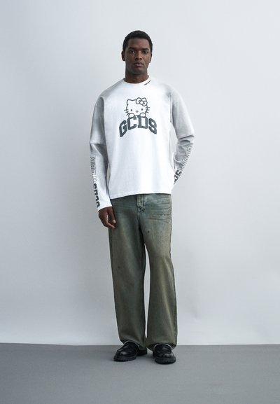 Άνδρας που στέκεται με γκρι τζιν παντελόνια φαρδιάς γραμμής, λευκό πουκάμισο μακρυμάνικο με Hello Kitty και κείμενο GCDS, και μαύρα παπούτσια σε απλό ανοιχτόχρωμο φόντο.