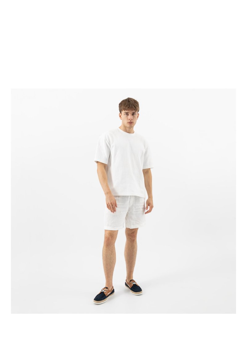 Un homme debout devant un fond blanc, portant un t-shirt blanc, un short blanc et des chaussures bateau bleu marine avec des lacets beige.