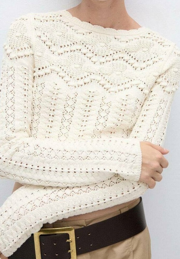 Pull blanc en maille à manches longues avec des motifs texturés, porté avec un pantalon beige et une large ceinture noire avec une grande boucle dorée.