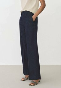 Breda byxor i mörk denim med hög midja och sidofickor, kombinerade med texturerade klacksandaler med ett ormtrycksmönster.