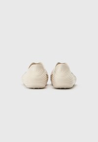 Beige slip-on shoes with a ridged design, rounded shape, and textured finish. Features a subtle logo on the heel.