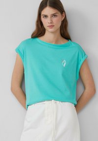 Turekowy bawełniany T-shirt z podwiniętymi krótkimi rękawami i okrągłym dekoltem. Posiada minimalistyczną białą grafikę na klatce piersiowej.