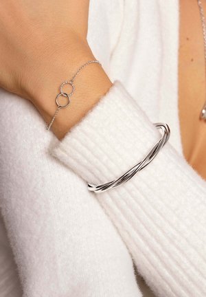 Bracelet en argent torsadé sur une main, associé à un délicat bracelet en argent avec deux cercles entrelacés, sur un pull blanc.