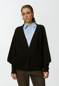 Crna rebrasta kardigan s širokim rukavima i prednjim džepovima, nosi se preko svijetloplavog košulje s kragnom. Materijal izgleda mekano i teksturirano.