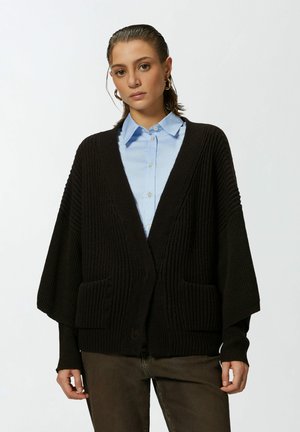 Cardigan noir à côtes avec des manches larges et des poches avant, porté par-dessus une chemise à col bleu clair. Le matériau semble doux et texturé.