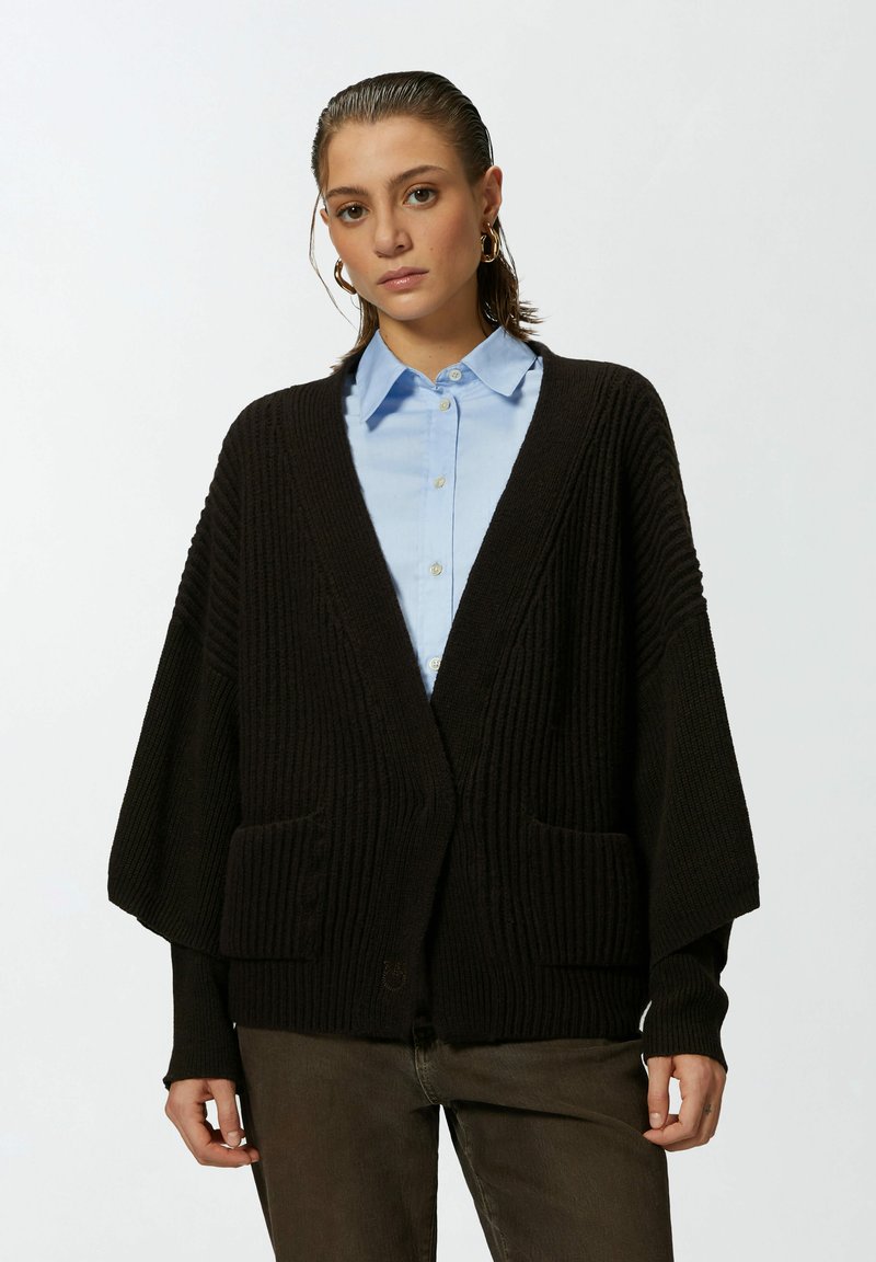 Crna rebrasta kardigan s širokim rukavima i prednjim džepovima, nosi se preko svijetloplavog košulje s kragnom. Materijal izgleda mekano i teksturirano.