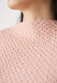 Strukturierter, weicher pinkfarbener Stoff mit einer gewellten, zerknitterten Oberfläche. Hoher Kragen mit subtiler Glanzwirkung, der sich auf das filigrane Muster und die Farbe konzentriert.