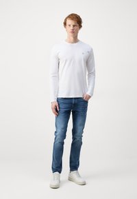 Hvid langærmet skjorte med rund halsudskæring, parret med lyseblå jeans og hvide sneakers, stående mod en ensfarvet baggrund.