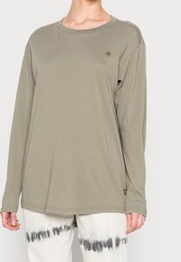 Person som bär en lös olivgrön långärmad skjorta och vita byxor med svarta tie-dye-mönster runt låren, som står mot en enfärgad bakgrund.