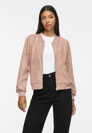 Femme portant une veste bomber en daim marron clair sur une chemise blanche et un pantalon noir, debout devant un fond blanc uni.