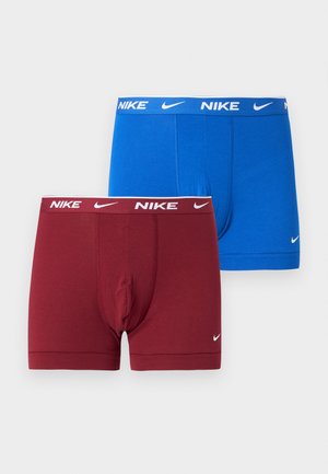 Uomo snello senza maglietta sta in piedi a piedi nudi su un tappeto, indossa boxer blu Nike, con una mano sul fianco e un'espressione neutra.