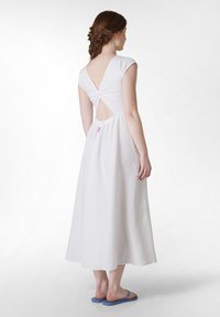 Rochie albă, până la podea, cu spate în formă de V și detalii răsucite. Material moale, mâneci scurte, talie strânsă, purtată cu flip-flops albastre.