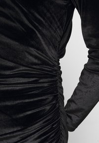 Robe longue en velours noir à manches longues avec des détails froncés le long de la couture latérale et une silhouette cintrée.