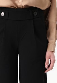 Svarta högmidjade byxor med veck, två dekorativa knappar och en slät tygtextur, ihopparade med en beige långärmad blus.