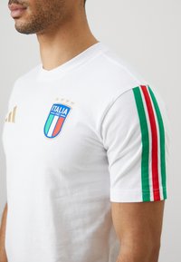 Λευκό βαμβακερό t-shirt με μπλε και πράσινο σήμα Ιταλίας, χρυσές λεπτομέρειες και πράσινες/κόκκινες ρίγες στις μανίκες. Μαλακή υφή.