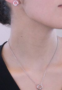 Collana d'argento con un pendente circolare e un piccolo accento, abbinata a orecchini a bottone circolari coordinati, indossata su una maglia scura.