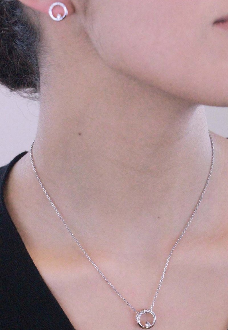 Collana d'argento con un pendente circolare e un piccolo accento, abbinata a orecchini a bottone circolari coordinati, indossata su una maglia scura.