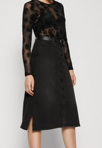 Kvinna som bär en svart genomskinlig långärmad topp med blommönster och en högmidjad svart knäppt midikjol med ett bälte.