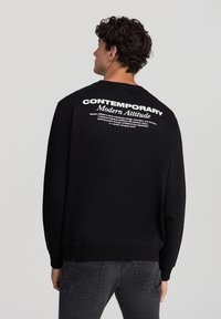 Felpa nera con scollo rotondo, caratterizzata da testo bianco sul retro. Il tessuto sembra morbido, con polsini e orlo a coste. Abbinata a jeans grigi.