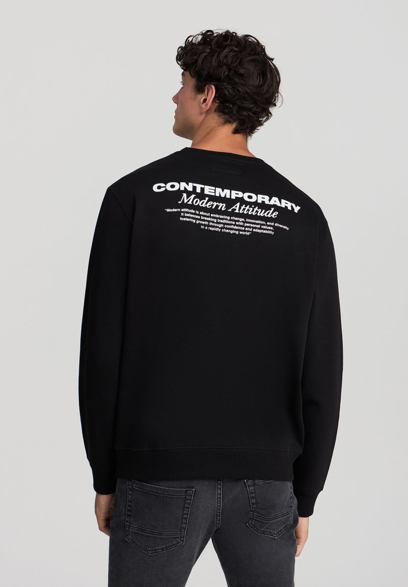 Felpa nera con scollo rotondo, caratterizzata da testo bianco sul retro. Il tessuto sembra morbido, con polsini e orlo a coste. Abbinata a jeans grigi.