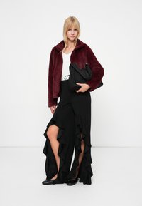 Noor wearing burgundia faux karusnahk jakk, valge topp, mustad volangpüksid jagatud sääreosaga, mustad korterjalatsid, hoides musta kottpussi.