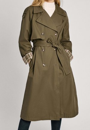 Femme portant un trench-coat kaki à double boutonnage avec des manches doublées de tissu à carreaux et une ceinture nouée, par-dessus un haut blanc à col montant.