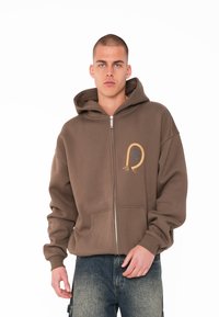 Brauner Reißverschluss-Hoodie mit großer Fronttasche, versehen mit einem goldenen Seil-Design und kreisförmigem Logo auf der linken Brust. Aus weichem Stoff und in lässiger Passform.