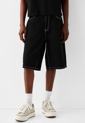 Person iført løse sorte knælange shorts med hvide syninger, hvide sokker, offwhite sneakers og en sort skjorte, stående indendørs.