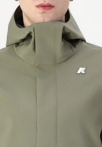 Jachetă impermeabilă de culoare verde măslinie, cu guler înalt, design minimalist și accent logo alb pe piept. Textură netedă cu cusături vizibile.