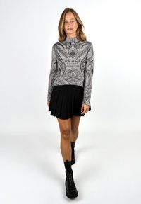 Woman walking forward wearing a gray patterned sweater with text, black pleated skirt, and black chunky ankle boots on white background.
