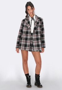 Donna con blazer a scacchi bianco e nero e pantaloni coordinati, camicia bianca con nastro nero, stivali neri alla caviglia, in piedi contro uno sfondo semplice.