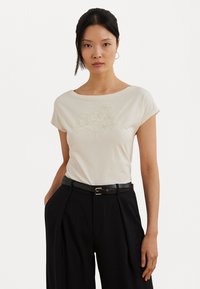 Cream-colored cotton t-shirt with a boat neckline and short sleeves, featuring floral embroidery on the front. Paired with black high-waisted trousers.