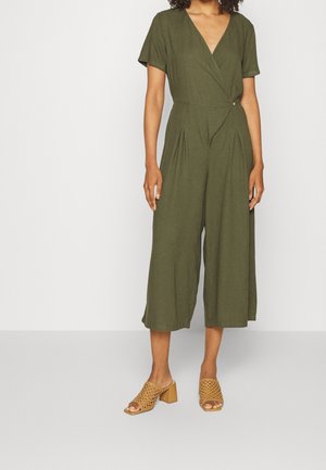 Mujer que lleva un mono de manga corta verde oliva con escote cruzado y piernas anchas y recortadas, combinado con sandalias de tacón bloque trenzadas en color tostado.