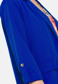 Erksinine bleiser siledast kangast, millel on nurgeline krae, rullitud varrukad kullast nupuga ning eesmise tasku detail.