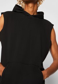 Black sleeveless hoodie made of soft fabric, featuring a large hood, front pocket, and raw edge detailing at the shoulders.