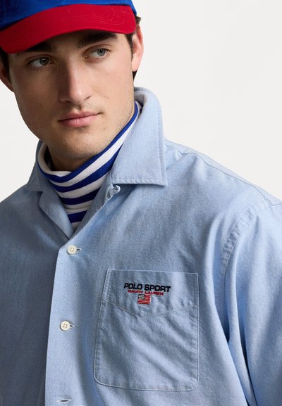 Light blue button-up shirt with a pocket, featuring a red "POLO SPORT" logo. Worn over a blue and white striped turtleneck.