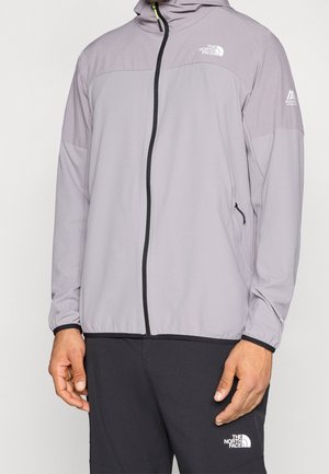 Mężczyzna noszący jasnoszarą kurtkę z kapturem i zamkiem oraz czarne spodnie, oba z widocznym logo The North Face.