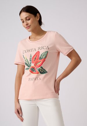 Femme portant une chemise rose clair avec un motif coloré de papaye et de feuilles et le texte "Costa Rica Papaya", associée à un pantalon blanc.