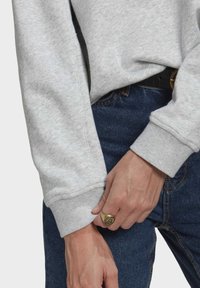 Grå sweatshirt med ribbade muddar, i kombination med mörkblå jeans. En guldring pryder en hand som greppar mudgarna.