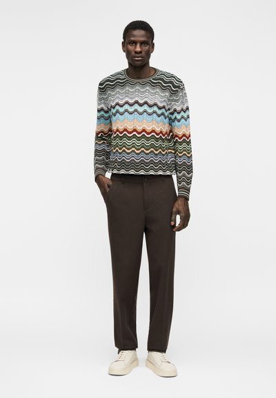 Άνδρας που φοράει πουλόβερ με πολύχρωμο ζιγκ-ζαγκ μοτίβο, σκούρο καφέ παντελόνι και κρεμ αθλητικά παπούτσια, στέκεται με το ένα χέρι στην τσέπη.