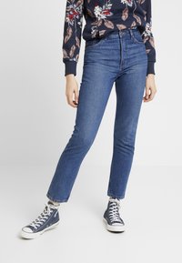 Jeans de mezclilla azul de cintura alta con un ajuste ajustado, estilo clásico de cinco bolsillos y dobladillo sin terminar. Combinados con zapatillas de lona oscura.