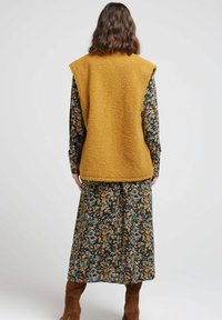 Un gilet sans manches jaune moutarde et duveteux porté sur une robe florale foncée à manches longues et longueur midi, associé à des bottes marron hautes jusqu'au genou.