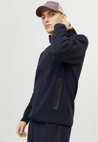 Marinefarbener Reißverschluss-Jacke mit seitlichen Taschen, aus dehnbarem Stoff. Strukturelle Akzente und ein erhöhter Kragen. Ergänzt durch eine beigefarbene Mütze.